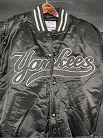 Front view of black satin New York Yankees jacket showing full 'Yankees' scripted logo and snap button front closure.