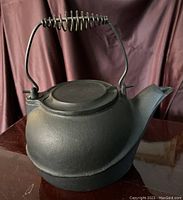 Front and side view of the antique large black cast iron kettle on a wooden surface, showing textured matte black finish, curved spout, hinged lid, and coil handle.