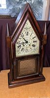 Front perspective of the antique cathedral mantle clock showing wooden pointed top, two side spires, Roman numeral dial with floral background, lattice bottom glass panel and dark wood case.