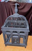 Photo of the antique or vintage birdcage on a dark cloth background, showing the cage front including wire dome top with ornate finial and the small lockable front door.