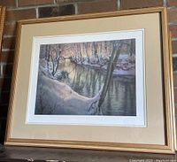 Framed vintage lithograph print showing winter river scene with snow and trees, matted and framed in a gold-tone wooden frame.