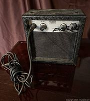 Front view of the First Act MA2039 miniature guitar amplifier showing black textured exterior, silver control panel with input, volume, gain, tone knobs, and black speaker grille.