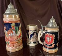 Three antique German beer steins displayed on a dark surface with brown backdrop, showing different heights and detailed relief motifs.