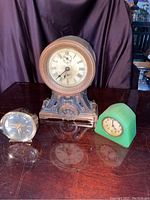 Photo showing all three clocks together: One large ornate antique clock with round face and Roman numerals, a smaller green plastic clock, and a smaller metal antique clock lying down.