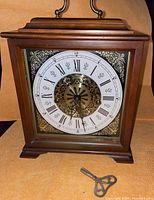 Front view of mantel clock showing wood case, brass handle, ornate dial and winding key in foreground