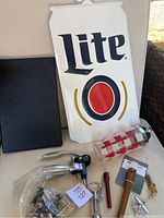 Tin Miller Lite sign leaning at back with black menu book, multiple wine openers, cocktail shaker, muddler, and pourers displayed on white surface.