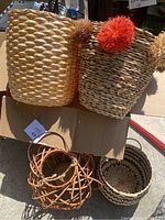 Box containing a collection of baskets: large woven baskets (one decorated with pom-poms), smaller woven baskets in natural and black tones, and a small metal frame basket.