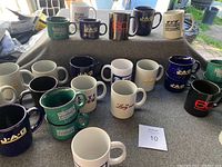 Photo showing a collection of 17 ceramic coffee mugs with various TV show and news logos placed on a gray cloth with outdoor background. Shows details of colors and logos on mugs.