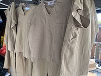 Several khaki correctional uniform tops and bottoms hanging from a rack showing front and side views. V-neck shirts with chest pockets visible.