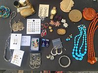 Full overview of costume jewelry collection laid out on black surface showing earrings, necklaces, compacts, boxes, and a wristwatch