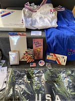 Lot contents including vintage white Olympics shirt, blue shorts, playing cards, pins, keychains, and collector plate boxed