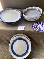 Six white ceramic plates with blue rims and four matching bowls arranged on a surface along with one decorative blue floral plate.