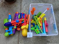 Assorted water soakers and foam water squirters placed on ground and in a clear plastic bin