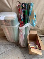 Photo showing multiple rolls of wrapping paper including holiday-themed ones, placed upright beside a translucent plastic Rubbermaid container and a cardboard box with ribbons.