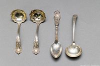 Four sterling silver spoons laid out flat showing detail of ornate handles and bowl shapes, including pierced decoration on two spoons.