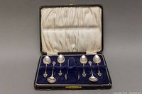 Seven-piece sterling silver set of espresso spoons and sugar tongs arranged in a fitted box with dark blue velvet interior and satin lining.