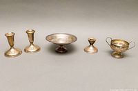 Wide view showing set of five silver items: two tall candleholders, one small pedestal bowl, one smaller candleholder, and one two-handled bowl. Items show tarnishing and dents.