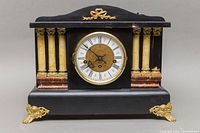 Front view of black mantel clock with four decorative columns on each side and gold ornamentation. Clock face visible with Roman numerals.