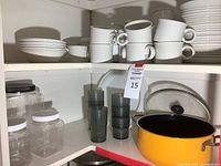 Shelves with stacked white china plates and mugs, plastic canisters, and a bright yellow Dutch oven with glass lid.