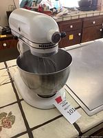 White KitchenAid stand mixer on countertop with stainless steel mixing bowl and wire whisk attachment.