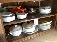 Wooden shelving unit with top and bottom shelves holding multiple cookware pieces including orange and cream Le Creuset items, stainless steel pan and white dishes