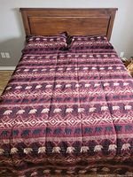 Photo of a bed dressed with a patterned duvet cover and two matching pillow shams. The design features repeating rows of black and cream bear silhouettes and geometric shapes in burgundy and red tones.
