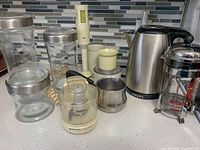 Photo showing nine kitchen items including Krups kettle, Bodum French press, Braun immersion blender with chopping attachment, Black & Decker Handy Chopper, small saucepan, and three glass containers with metal lids. Items arranged on countertop with tiled backsplash.