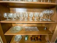 Top and middle shelves showing variety of stemmed wine glasses, goblets, plates, a rectangular cut crystal tray, and wooden candle holders below.