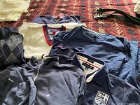 Tommy Hilfiger shirts and sweaters arranged on a bed, showing the front of each in natural light. Some have the Tommy Hilfiger logos and multi-color trim.
