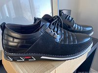 Side profile of one black Gatsby men's shoe, showing stitched upper with smooth and suede-like panels and patterned rubber sole with white and red accents.