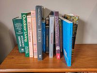 Photo shows a collection of various hardcover and softcover books standing vertically on a wooden surface, including titles related to outdoor living, Leeds & Lansdowne history, and woodworking guides.