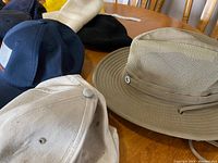 Several hats displaying variety and styles including beige outdoorsman hat, various baseball caps in navy and black, shown on wooden table with chair backs