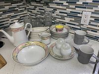 Kitchen counter display showing teapot, clear glasses, plates, bowls, mugs, creamer and salt and pepper shakers with tile backsplash and electrical outlet