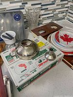 View of lot on kitchen counter showing canning jars box, funnel, plastic cutlery, paper plates, wine glasses, wooden trays, glass vase, and small fondue set