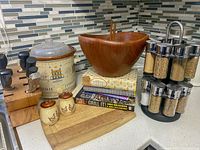 Photo showing wooden salad bowl with tongs, two wooden bowls, crockpot, spice rack, cutting board, knife block with knives, salt and pepper shaker, and cookbooks.