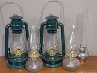 Photo showing five oil lamps: two green Dietz lanterns, two round glass base lamps, one hammered silver-like base lamp, and small bottle of oil