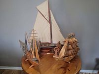 Photo showing four wooden model boats arranged on a round wooden table: a large sailboat with beige sail, a multiple-masted sailboat, a smaller boat with fishing net, and a wooden fisherman statue.