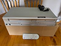 Silver Sylvania SV2000 combo VHS and DVD player placed on cardboard box with white remote control and power cord on top.