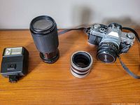 Canon AT-1 camera body with Canon lens attached, zoom lens, external flash, and camera strap laid out on wooden surface