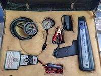 Open hard black case showing timing light with pistol grip, vacuum gauge, compression tester, test meter, all positioned in foam cutouts.