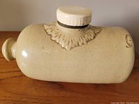 Side view of vintage ceramic hot water bottle/foot warmer showing screw cap and decorative floral pattern around neck.