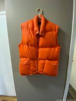 Front view of the bright orange puffer vest hung on white hanger, showing padded horizontal quilting and front zipper.