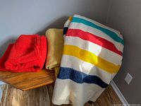 Three folded wool blankets on wooden surface including striped Trapper Point blanket, solid red and solid yellow blankets.