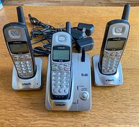 Photo showing three VTech cordless telephone handsets with silver base stations and antennas, placed on wooden surface with power adapters beside them.