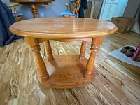 Angle view of the solid wooden oval side table showing top surface, legs and bottom shelf on wood flooring