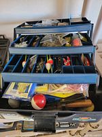 View of the open fishing tackle box showing multiple compartments filled with fishing lures, bobbers, and other tackle equipment