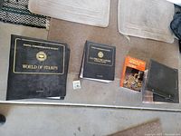 Four large items on a carpeted surface including two Postal Commemorative Society albums, one book on coins, and a small box of stamps.