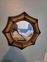 Photo showing the round mirror with sunburst frame mounted on a light gray or blue wall, reflecting part of a room with a window and bedding.