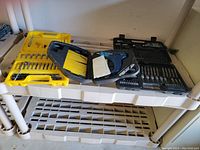 Three plastic cases containing tools on a table, including two drill bit sets and one cordless drill kit.