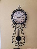 Full view of ornate black metal frame wall clock with pendulum, round clock face with Roman numerals and black decorative hands.